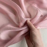 Une main soulève une Mousseline de soie de mûrier de luxe rose et transparente de Longan Craft France, mettant en valeur sa texture naturelle, légère et pure sur un fond clair.