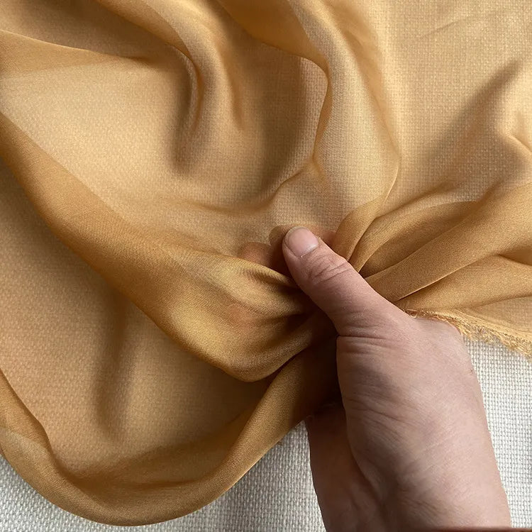 Une main tient une section de la Mousseline de soie de mûrier de luxe au mètre de Longan Craft France, montrant sa texture diaphane et douce - idéale pour les vêtements de luxe - sur un fond neutre.