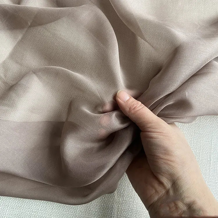 Une main tient un morceau de Longan Craft France Mousseline de soie de mûrier de luxe au mètre. Cette mousseline de soie naturelle, diaphane et marron clair est lisse et translucide, idéale pour les vêtements de luxe.