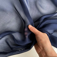 Une main fronce la Mousseline de soie de mûrier de luxe au mètre bleu foncé de Longan Craft France, mettant en valeur la translucidité naturelle du tissu et sa texture douce et légère sur un fond clair.