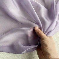 Une main tient la luxueuse mousseline de soie de mûrier violet clair de Longan Craft France, un tissu de robe naturel, léger et transparent, sur un fond texturé blanc cassé.