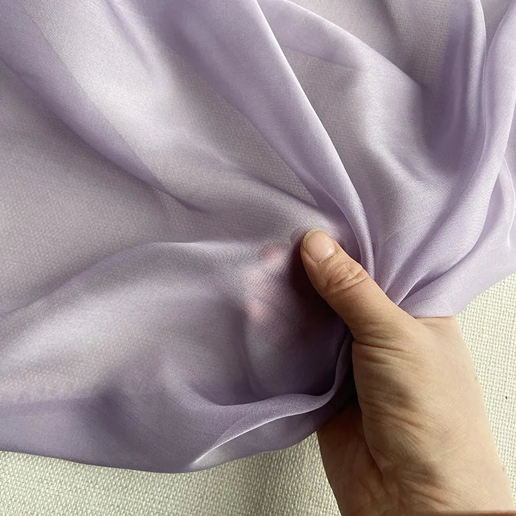 Une main tient la luxueuse mousseline de soie de mûrier violet clair de Longan Craft France, un tissu de robe naturel, léger et transparent, sur un fond texturé blanc cassé.