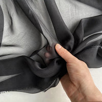 La mousseline de soie de mûrier de luxe au mètre de Longan Craft France est délicatement froncée par une main, révélant la texture naturelle, légère et transparente de ce tissu pur sur un fond clair.