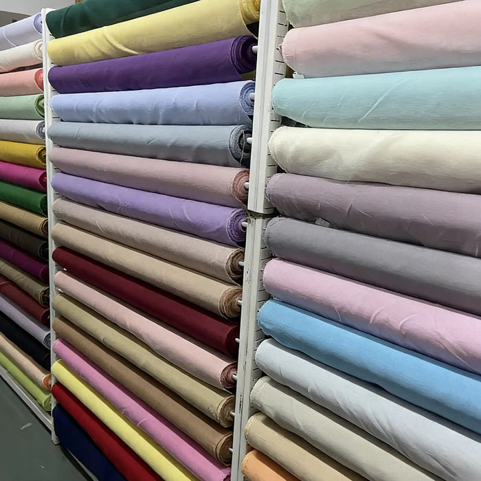 Les étagères présentent le Tissu soie et lin 14 mommes au mètre de Longan Craft France - organza de soie de mûrier en violet, bleu, rose, vert, beige et jaune vibrant - organisé horizontalement sur des étagères métalliques dans le magasin.