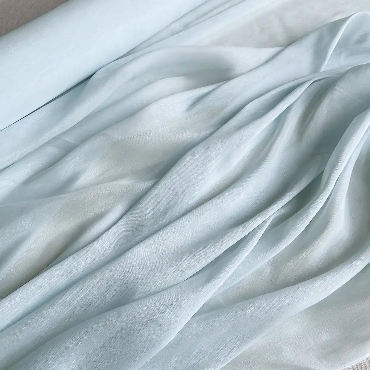 Le Tissu soie et lin 14 mamans au mètre de Longan Craft France est un organza de soie de mûrier semi-transparent et léger, présenté partiellement déroulé pour révéler ses plis doux et sa texture bleu pâle luxueuse, parfaite pour l'été.