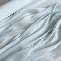 Le Tissu soie et lin 14 mamans au mètre de Longan Craft France est un organza de soie de mûrier semi-transparent et léger, présenté partiellement déroulé pour révéler ses plis doux et sa texture bleu pâle luxueuse, parfaite pour l'été.