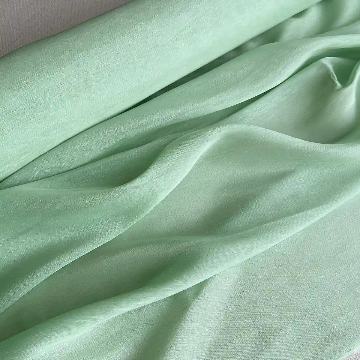 Un gros plan sur Tissu soie et lin 14 mommes de Longan Craft France révèle sa texture douce et lisse en organza de soie de mûrier vert clair, avec des plis doux et des lignes ondulées - un mélange de soie et de lin idéal pour l'été.