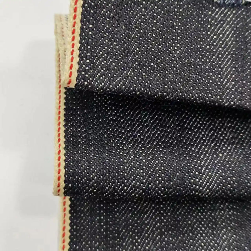 Gros plan sur le Tissu en jean selvedge 16,5 oz de Longan Craft France : denim épais bleu foncé, 100% coton, avec une armure diagonale blanche et un bord beige avec une bande rouge. La texture et les détails de qualité supérieure sont clairement visibles.
