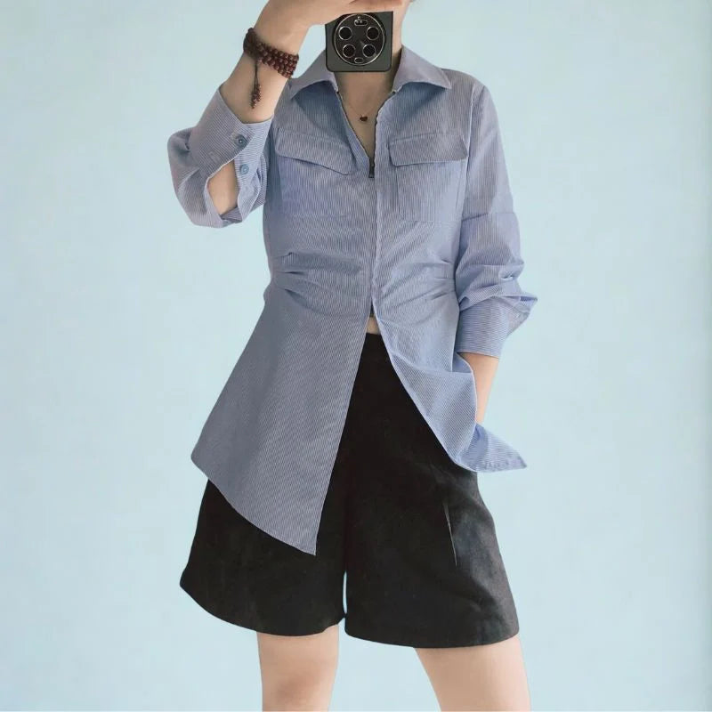 Une personne se tient debout sur un fond bleu clair, portant le Patron de couture chemisier zippé pour femme de Longan Craft France avec un short noir. Son visage est caché par un téléphone, une main dans la poche. Patron au format PDF à télécharger gratuitement.