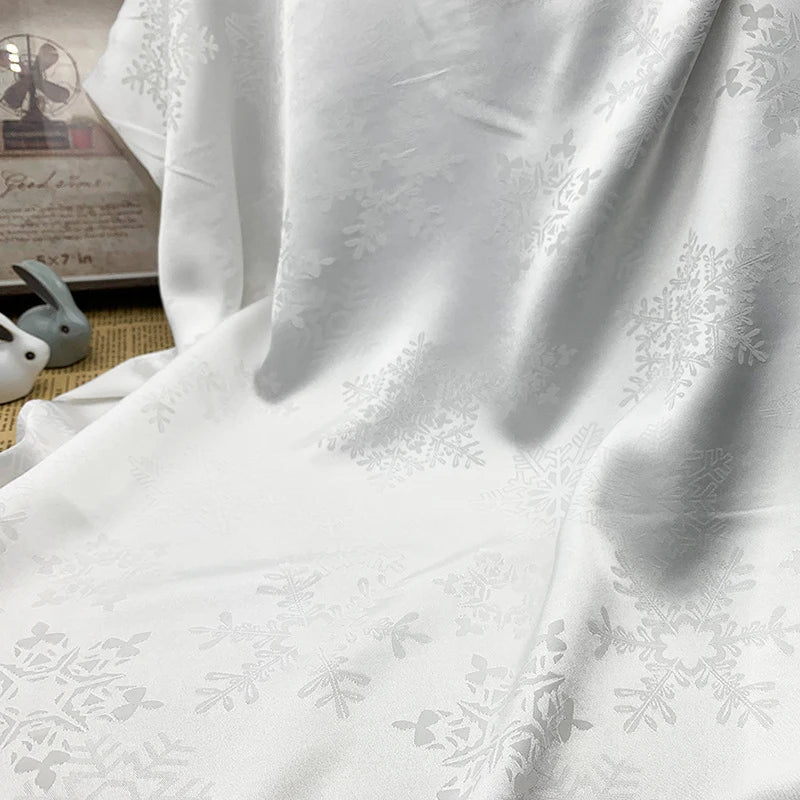 Un gros plan du tissu satin jacquard motif flocon au mètre de Longan Craft France, un tissu lisse en polyester blanc pour robes, montre son motif subtil de flocon de neige et sa texture soulignée par des ombres douces. Les objets en arrière-plan sont légèrement flous.
