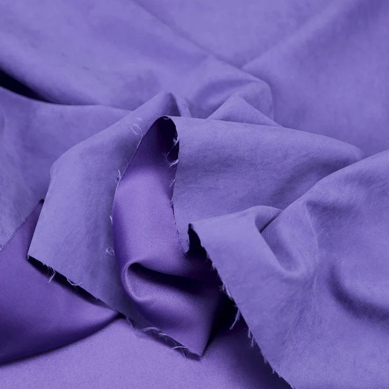 Un gros plan du microsuède polyester multicolore au mètre de Longan Craft France montre sa douce texture peau de pêche brossée, avec des bords dentelés et des plis doux sur un fond violet assorti - idéal pour la création de vêtements.