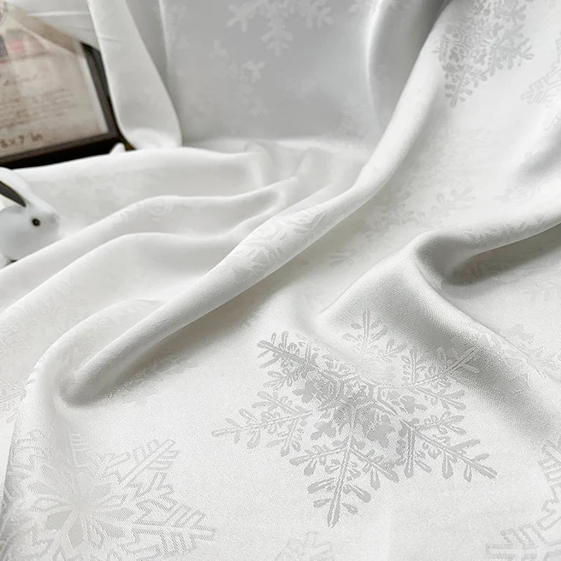 Le tissu satin jacquard motif flocon au mètre de Longan Craft France, un textile polyester blanc lisse pour robes, est drapé pour montrer des plis doux et des ombres, avec un arrière-plan flou et une petite souris blanche.