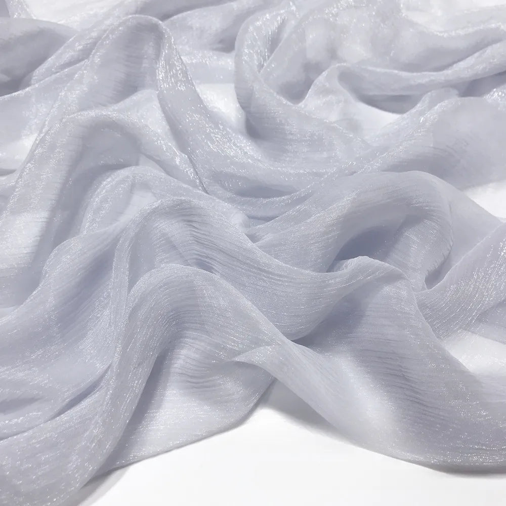 Gros plan sur le tissu mousseline plissé brillant au mètre de Longan Craft France - un tissu polyester gris clair, transparent et brillant - drapé en vagues douces sur une surface plane.