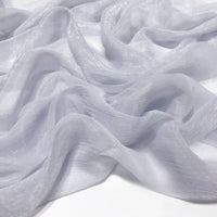 Gros plan sur le tissu mousseline plissé brillant au mètre de Longan Craft France - un tissu polyester gris clair, transparent et brillant - drapé en vagues douces sur une surface plane.