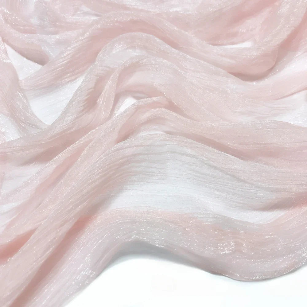 Gros plan sur le tissu mousseline plissé brillant au mètre de Longan Craft France, un polyester léger rose pâle au lustre subtil et aux plis ondulés, drapé délicatement sur un fond blanc.
