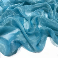 Un gros plan de Tissu mousseline plissé brillant au mètre de Longan Craft France montre un tissu de polyester translucide, bleu clair et brillant, drapé et plié sur une surface blanche.