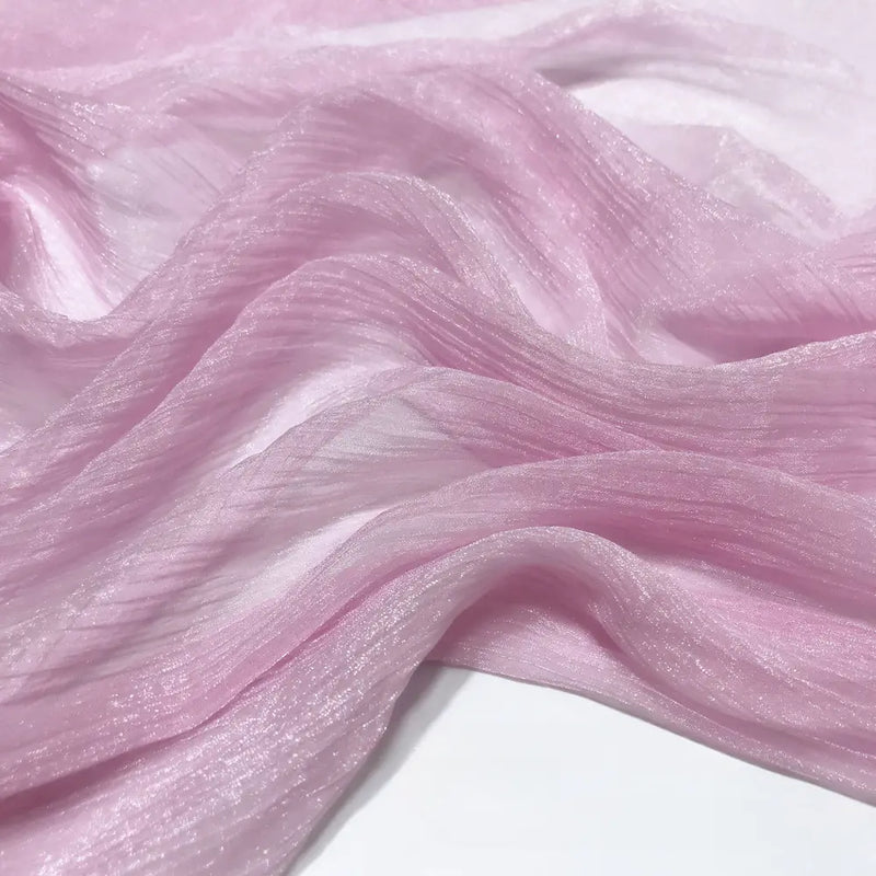 Gros plan sur le Tissu mousseline plissé brillant au mètre de Longan Craft France, une mousseline de polyester rose pâle, transparente et chatoyante, à la texture froissée, drapée en plis souples sur une surface blanche.