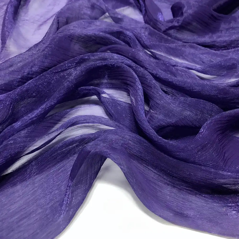 Gros plan sur le tissu mousseline plissé brillant au mètre de Longan Craft France dans un coloris violet profond, mettant en valeur ses plis légers, chatoyants et translucides sur une surface blanche. Cette mousseline de polyester froissée est douce et fluide.