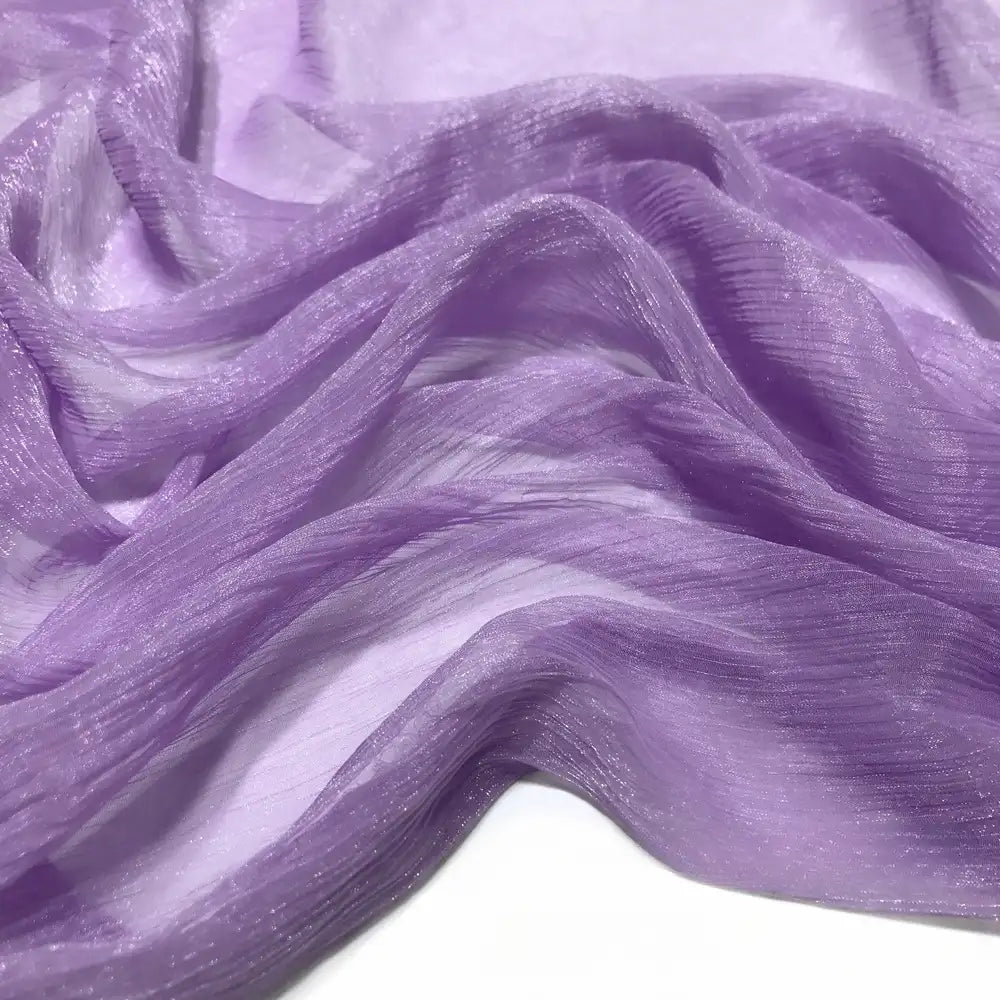 Gros plan sur le Tissu mousseline plissé brillant au mètre de Longan Craft France, un tissu de polyester transparent, léger et chatoyant dans les tons lavande, délicatement drapé en plis doux sur une surface blanche.