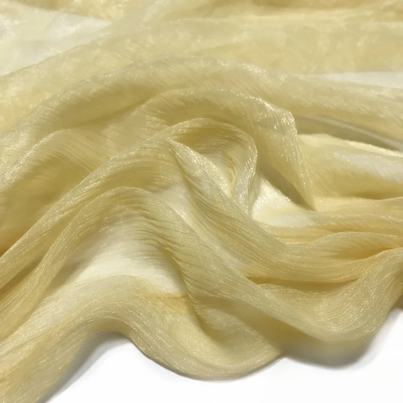 Un gros plan du tissu mousseline plissé brillant au mètre de Longan Craft France montre sa texture polyester jaune clair, semi-transparente, chatoyante et légèrement froissée, étalée en plis souples sur une surface blanche.