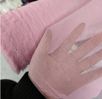 Une main présente du Tissu mousseline rose plissé brillant au mètre de Longan Craft France ; sa transparence et sa texture froissée sont visibles, avec un rouleau du même tissu polyester léger à côté, sur un fond doucement flou.