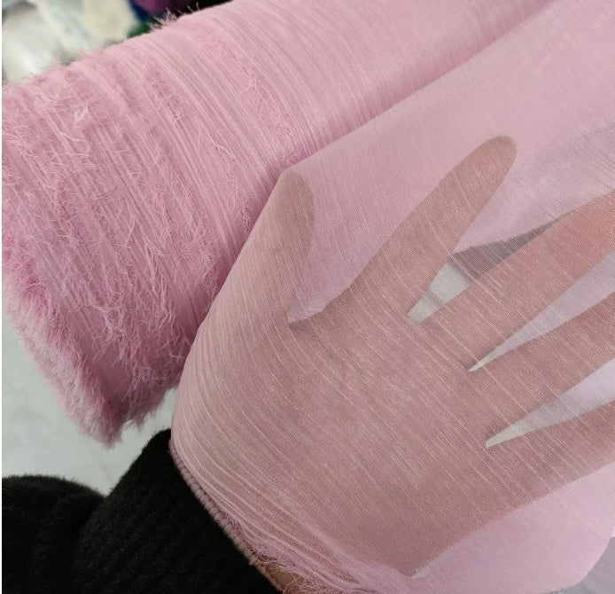 Une main présente du Tissu mousseline rose plissé brillant au mètre de Longan Craft France ; sa transparence et sa texture froissée sont visibles, avec un rouleau du même tissu polyester léger à côté, sur un fond doucement flou.