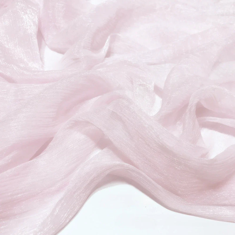 Un gros plan du tissu mousseline plissé brillant au mètre de Longan Craft France montre le tissu polyester rose pâle aux plis délicats, à la finition translucide et à la brillance subtile, drapé sur une surface blanche.