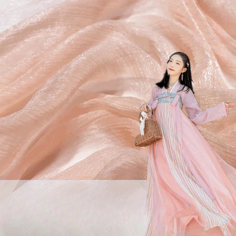 Une femme vêtue d'une robe rose fluide se promène avec un sac à main en osier, sur fond de Tissu mousseline plissé brillant au mètre de Longan Craft France, ressemblant à un délicat tissu de robe de soirée.