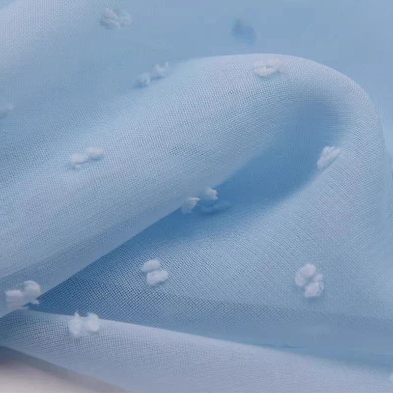 Gros plan sur Mousseline à plumetis au mètre de Longan Craft France : tissu jacquard doux avec des points brodés multicolores sur une surface en polyester bleu clair, légèrement translucide. Les plis doux ajoutent une profondeur élégante et ombragée.