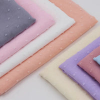 Les piles superposées de la Mousseline à plumetis au mètre de Longan Craft France affichent de douces teintes pastel - gris, rose, blanc, pêche, jaune, lavande et bleu clair - avec une délicate texture jacquard en pointillés.
