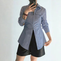 Personne prenant un selfie dans un miroir, vêtue d'une chemise à manches longues bleu clair inspirée du Patron de couture chemisier zippé pour femme de Longan Craft France, stylisée de manière asymétrique avec un short noir. L'arrière-plan est uni et clair.