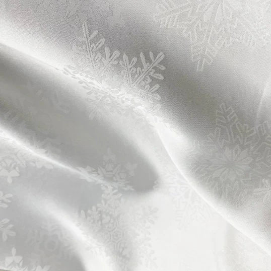 Gros plan sur le tissu satin jacquard motif flocon au mètre de Longan Craft France : tissu satin jacquard de polyester blanc, subtilement brillant avec un motif flocon de neige, légèrement plié pour montrer sa texture lisse - idéal pour les robes.