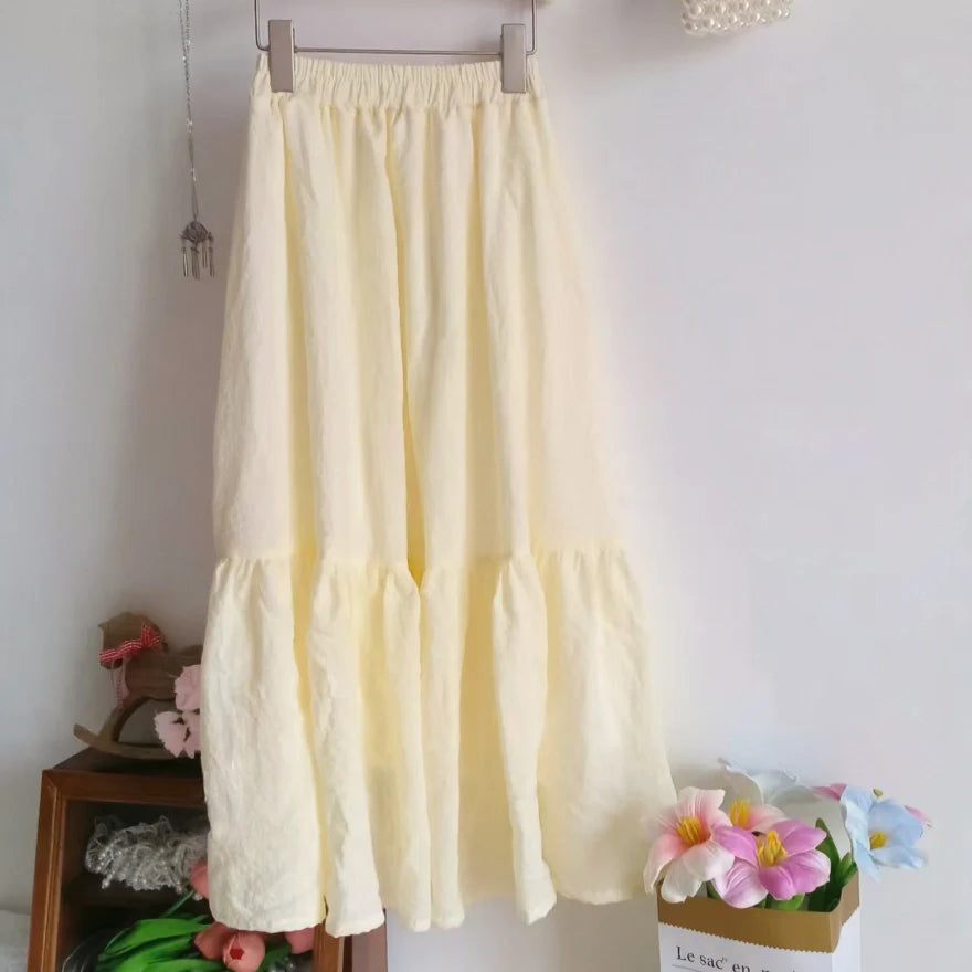 Un "Jupe midi plissée femme" jaune clair de Longan Craft France est suspendu à un cintre contre un mur blanc, avec des objets décoratifs et des fleurs artificielles en arrière-plan. Patron de couture gratuit au format PDF à télécharger.