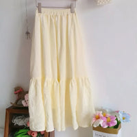 Un "Jupe midi plissée femme" jaune clair de Longan Craft France est suspendu à un cintre contre un mur blanc, avec des objets décoratifs et des fleurs artificielles en arrière-plan. Patron de couture gratuit au format PDF à télécharger.