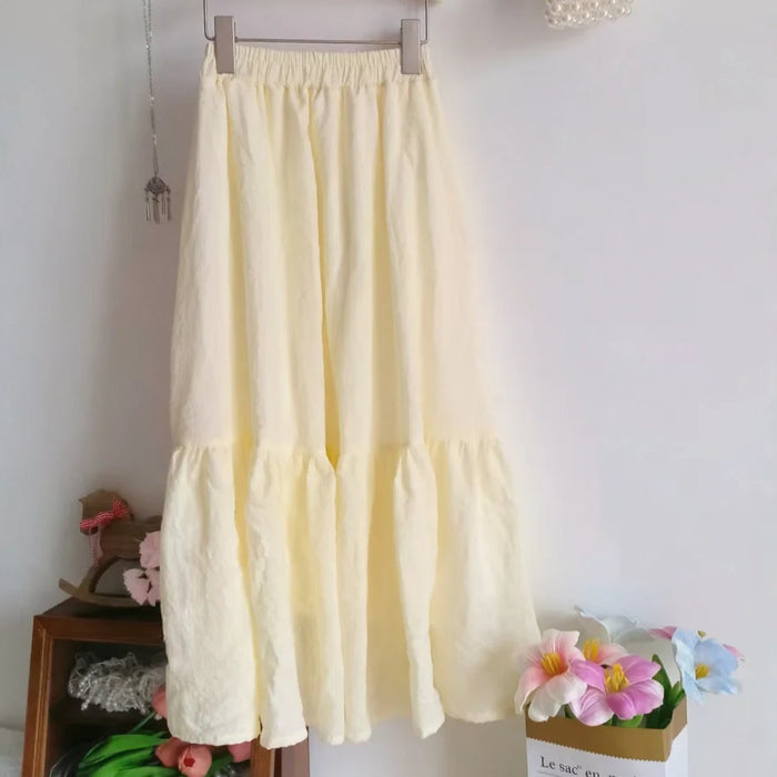 Un "Jupe midi plissée femme" jaune clair de Longan Craft France est suspendu à un cintre contre un mur blanc, avec des objets décoratifs et des fleurs artificielles en arrière-plan. Patron de couture gratuit au format PDF à télécharger.