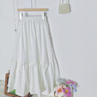 Une Jupe midi plissée femme de Longan Craft France (patron de couture PDF gratuit à télécharger) est suspendue à un cintre en bois contre un mur blanc, avec un sac à main en perles au-dessus et des fleurs artificielles colorées en bas à droite.