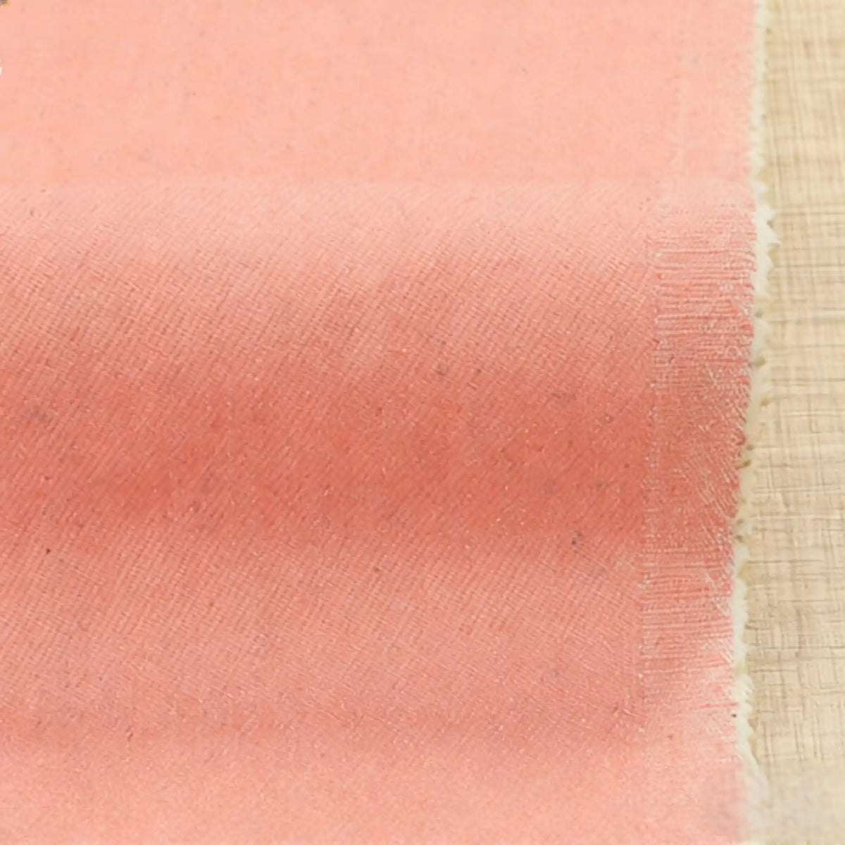 Gros plan sur le tissu Tissu denim extensible 9,5 oz de Longan Craft France - un tissu doux en coton-polyester de couleur pêche avec un tissage visible et des bords rosés, idéal pour les jeans - présenté sur une surface en bois clair.