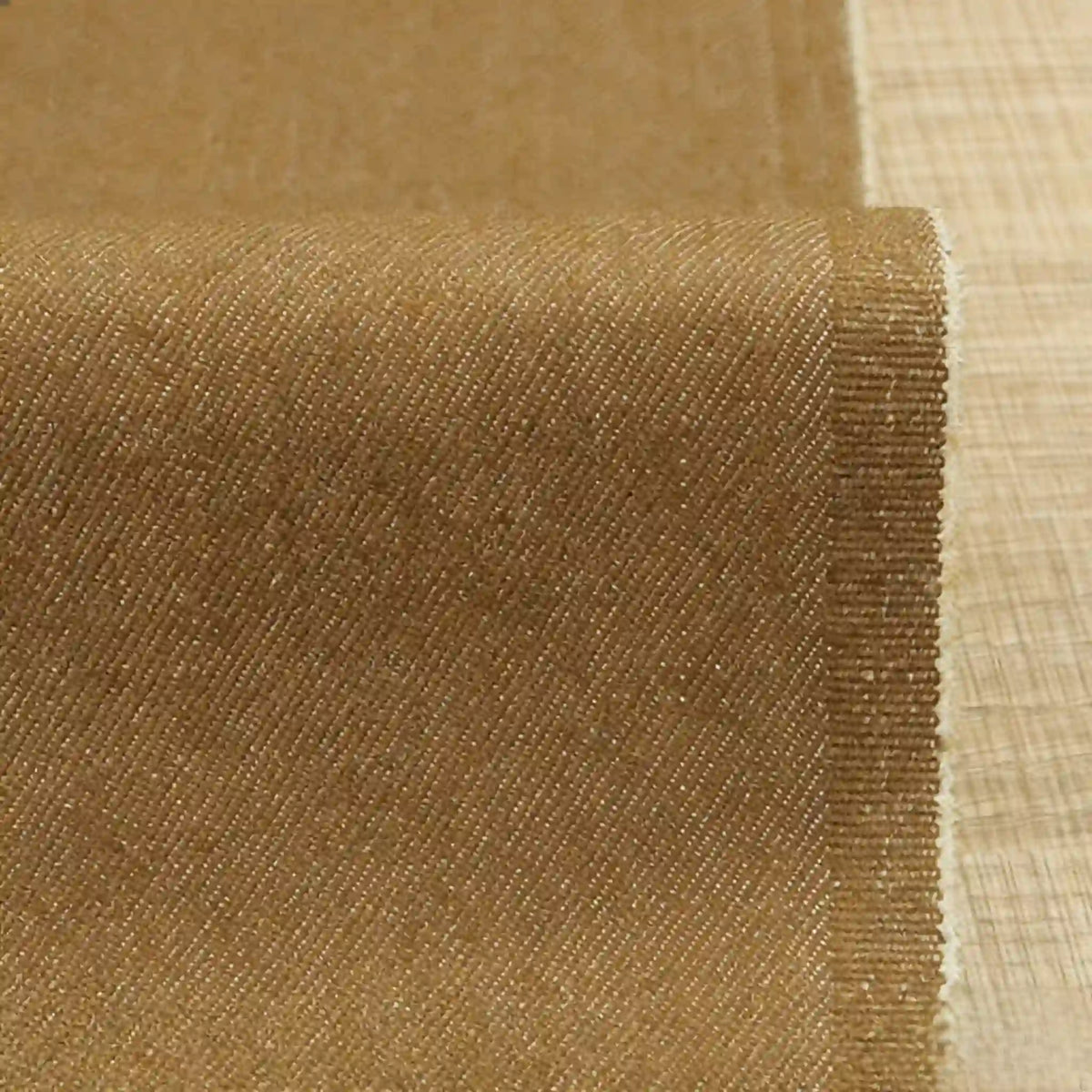 Un gros plan du Tissu denim extensible 9,5 oz au mètre de Longan Craft France montre son tissage lisse en coton-polyester idéal pour les jeans, contrastant avec la texture rugueuse de la toile de jute brune, le denim étant partiellement déroulé sur une surface plane.
