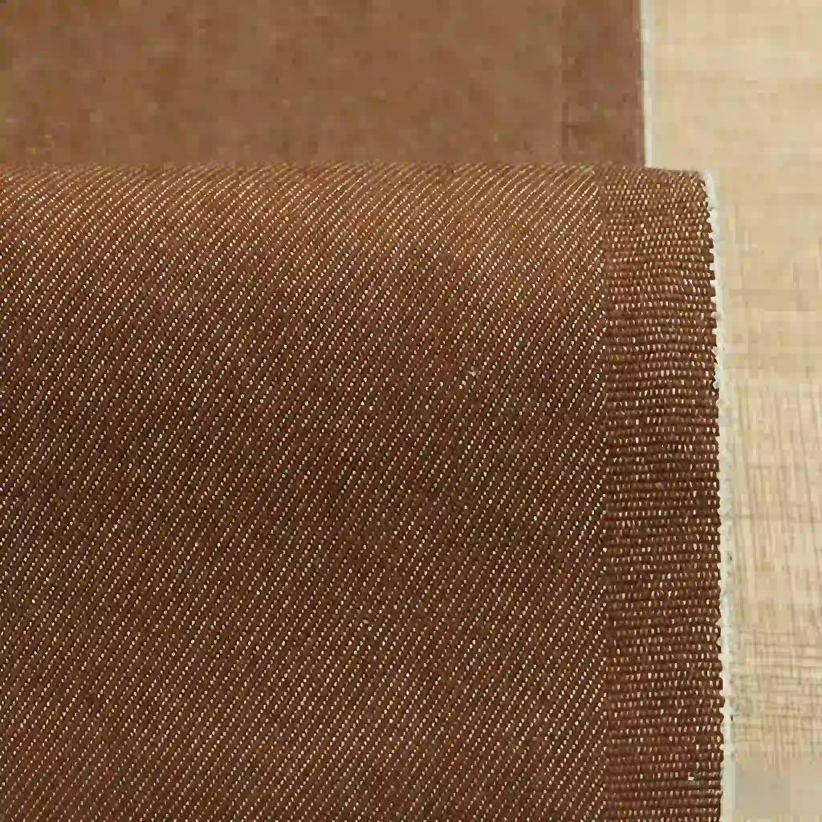 Gros plan sur le Tissu denim extensible 9,5 oz au mètre de Longan Craft France, un tissu brun en coton-polyester avec un tissage diagonal subtil, idéal pour les jeans. Le bord du tissu est net et droit, partiellement déroulé sur une surface plane.