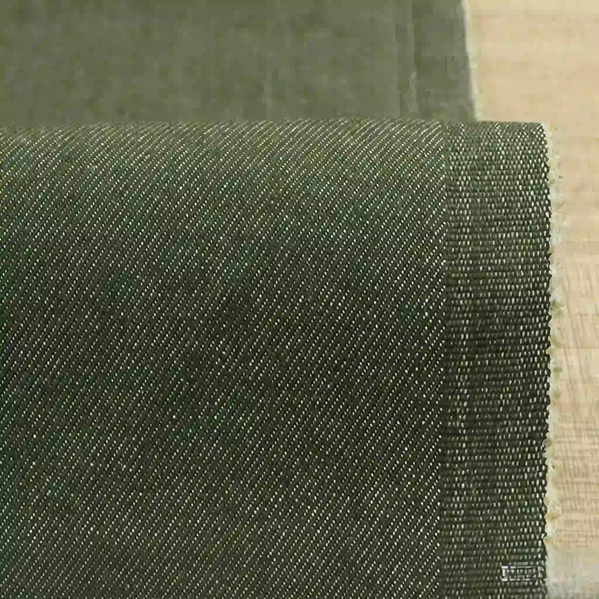 Gros plan sur un denim extensible Tissu vert foncé 9,5 oz de Longan Craft France, montrant sa fine trame diagonale et son bord légèrement effiloché, posé sur une surface en bois clair-idéal denim coton-polyester coloré pour jeans.