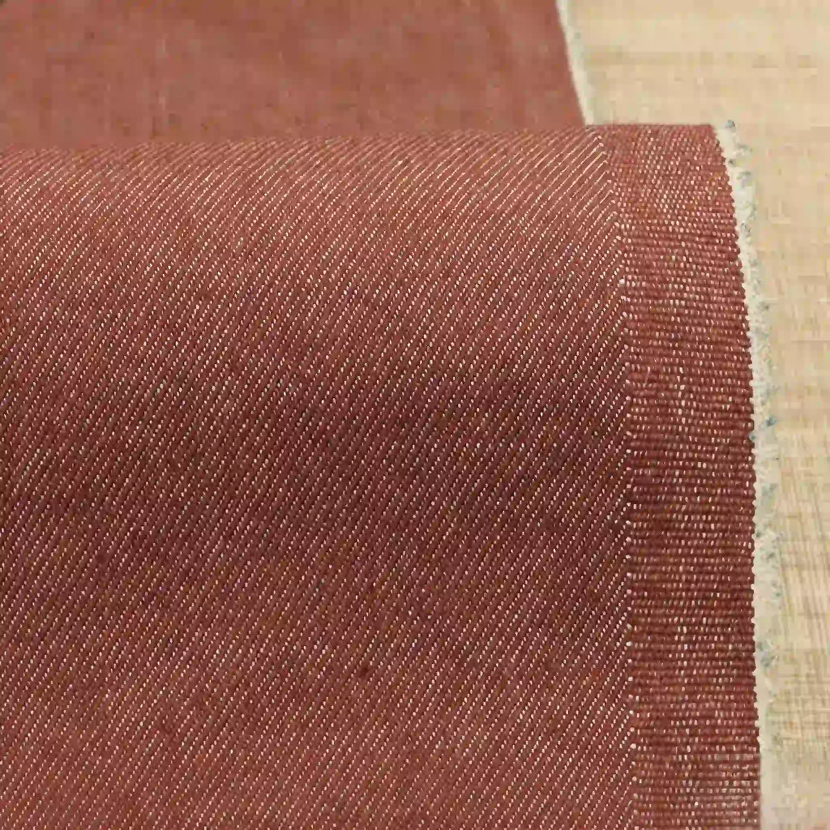 Un gros plan du Tissu denim extensible 9,5 oz au mètre de Longan Craft France montre son tissage coton-polyester brun-rouge avec un motif diagonal subtil sur une surface claire ; le bord du tissu apparaît à droite.