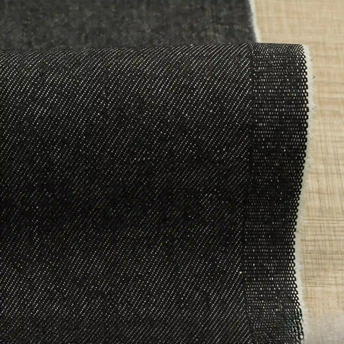 Un gros plan montre le Tissu denim extensible 9,5 oz au mètre de Longan Craft France - un denim sombre en coton-polyester tissé en diagonale avec un bord fini, parfait pour les jeans, présenté sur une surface texturée marron clair.