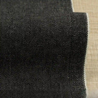 Un gros plan montre le Tissu denim extensible 9,5 oz au mètre de Longan Craft France - un denim sombre en coton-polyester tissé en diagonale avec un bord fini, parfait pour les jeans, présenté sur une surface texturée marron clair.