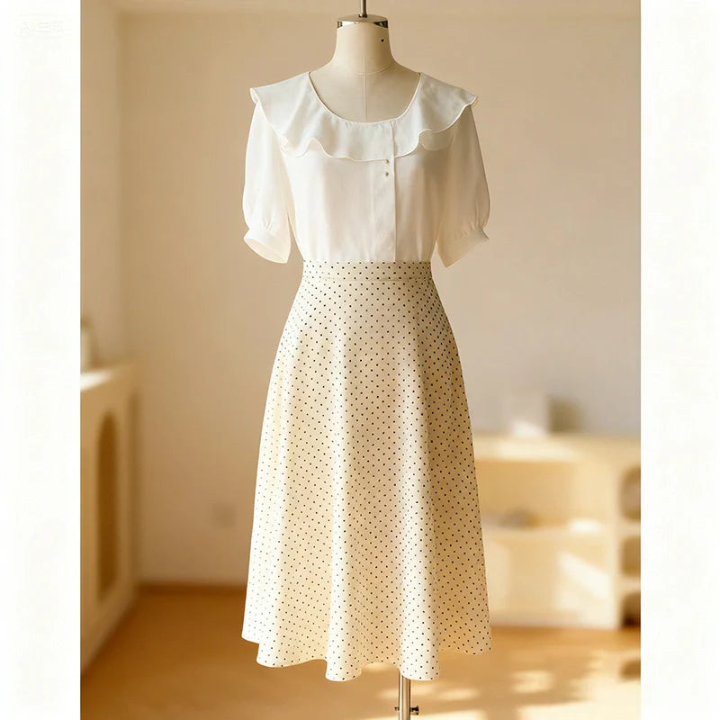 Un mannequin présente un chemisier blanc à col volanté et à manches courtes, associé au Patron de jupe midi trapèze pour femme de Longan Craft France - de couleur crème, à taille haute et à pois. L'arrière-plan est doucement éclairé et minimaliste.