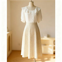 Un mannequin présente un chemisier blanc à col volanté et à manches courtes, associé au Patron de jupe midi trapèze pour femme de Longan Craft France - de couleur crème, à taille haute et à pois. L'arrière-plan est doucement éclairé et minimaliste.