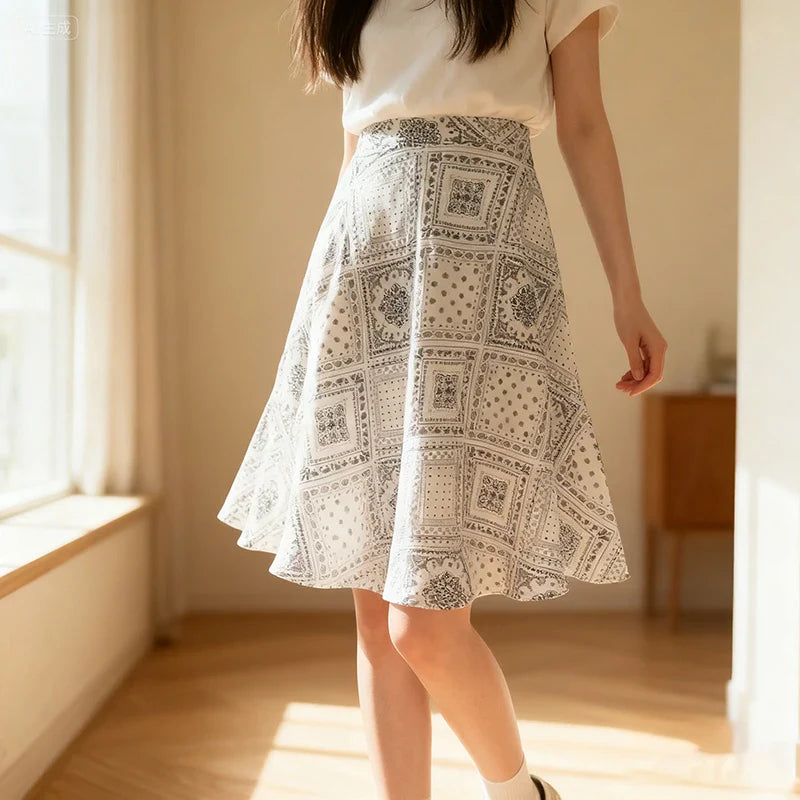 Une femme porte un haut blanc et une jupe midi fleurie grise à taille haute. Découvrez le "Patron de jupe midi trapèze pour femme | PDF gratuit à télécharger" de Longan Craft France - parfait pour coudre votre propre jupe tendance. La lumière du soleil envahit la pièce.