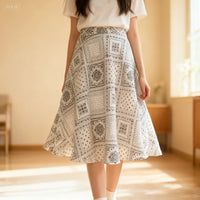 Une personne se tient à l'intérieur sur un sol en bois, à la lumière naturelle, vêtue d'un t-shirt blanc et d'une jupe midi trapèze à taille haute en Patron de jupe midi trapèze pour femme PDF de Longan Craft France, avec un motif gris et blanc.