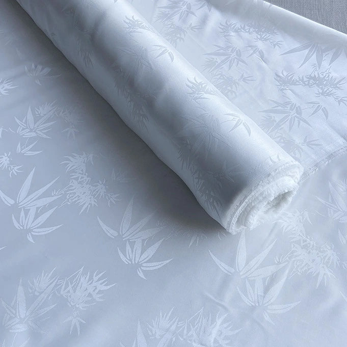 Un rouleau de tissu soie de mûrier et bambou imprimé jacquard au mètre de Longan Craft France est partiellement déroulé, montrant un motif de feuille subtil et brillant tissé dans le doux mélange de soie et de rayonne multicolore pour un aspect texturé et élégant.