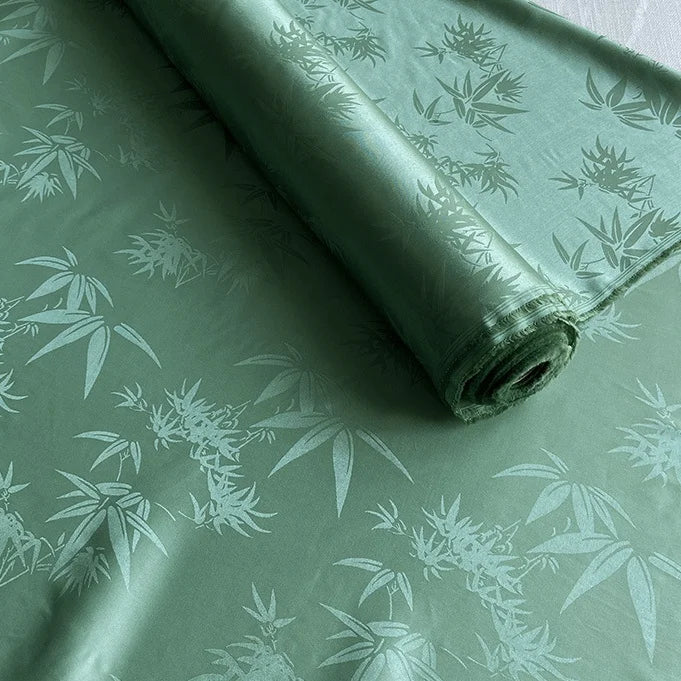 Un tissu Longan Craft France soie de mûrier et bambou imprimé jacquard au mètre, partiellement déroulé, met en valeur son subtil motif de feuilles de bambou et son lustre raffiné, parfait comme tissu de doublure multicolore doux et confortable en soie et rayonne.