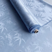 Un rouleau de Tissu soie de mûrier et bambou imprimé jacquard au mètre de Longan Craft France, un tissu de doublure en soie et rayonne multicolore, doux et confortable, est partiellement déroulé, mettant en valeur son subtil motif de feuilles et de branches de bambou.